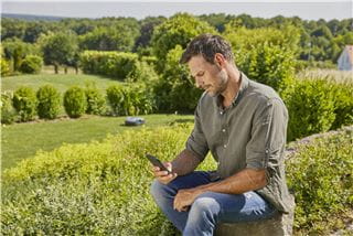 A man sitting in his garden looking at his smartphone with a gardena robotic lawnmower in the background